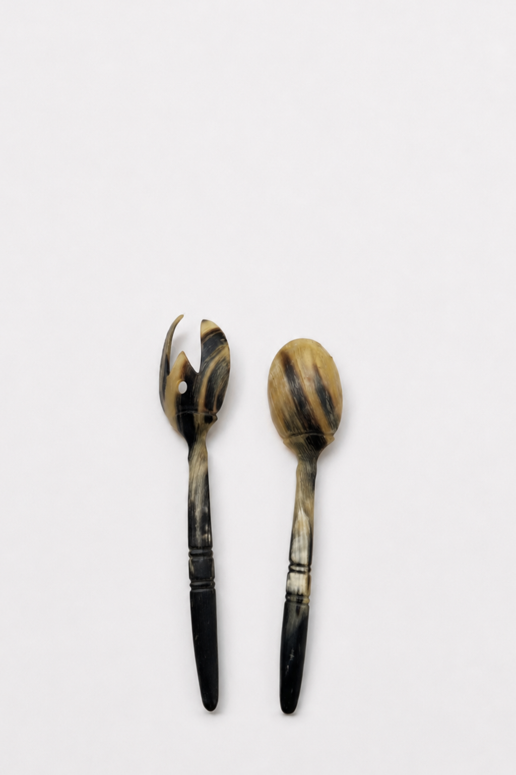 Two decorative serving utensils, a salad server on the left and a spoon on the right, featuring marbled black and tan handles with dark ferrules, set against a plain light gray background.